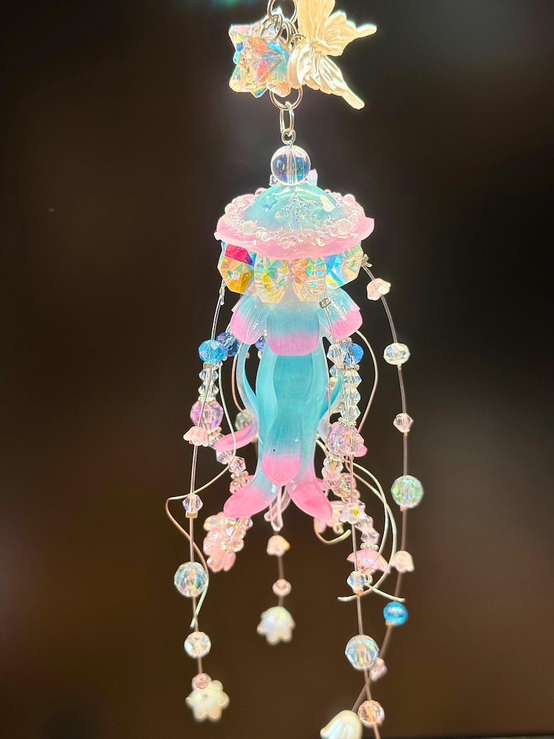 May include: A whimsical, handmade jellyfish ornament. The ornament features a light blue body with pink and blue accents, adorned with iridescent beads and dangling floral charms. A fairy-shaped charm and star-shaped charm are at the top.