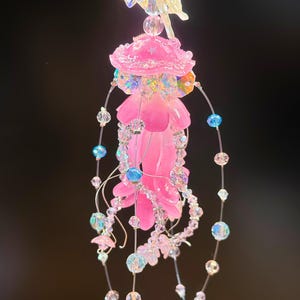 May include: A whimsical, pink and iridescent fairy-themed ornament. The ornament features a central pink flower shape, adorned with clear and iridescent beads, and a delicate fairy figure at the top. Strands of beads and small flowers dangle below.