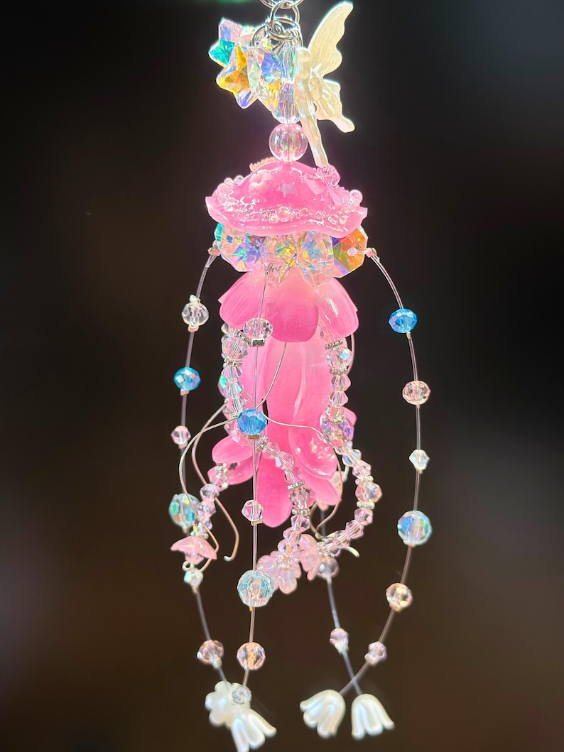 May include: A whimsical, pink and iridescent fairy-themed ornament. The ornament features a central pink flower shape, adorned with clear and iridescent beads, and a delicate fairy figure at the top. Strands of beads and small flowers dangle below.