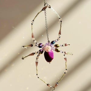 Könnte beinhalten: Ein dekoratives Spinnenornament aus Perlen. Es hat einen lila Körper und schillernde Beine. Die Spinne wird von einer silbernen Kette gehalten. Die Perlen sind in verschiedenen Lila- und Rosatönen gehalten.