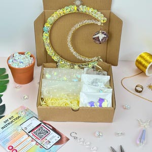 May include: A craft kit featuring a crystal crescent moon decoration. The kit contains craft materials like wire and crystals, and instructions. A small terracotta pot filled with crystals is also present, along with a smartphone displaying a QR code.