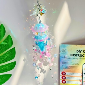 May include: A hanging jellyfish suncatcher with a blue, pink, and clear beaded design. The suncatcher has a jellyfish shape with a pink and blue top and a clear beaded body. A DIY kit instruction card is visible.