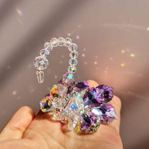 May include: A decorative crystal swan figurine with a curved neck and a flower-shaped body. The swan is made of clear and purple crystals, with iridescent accents. The figurine is held in a hand against a soft purple background.