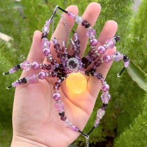 May include: A decorative spider ornament crafted from purple, black, and clear faceted beads, featuring a glowing yellow spherical abdomen. The spider has eight articulated legs made of strung beads and a central body with a silver-toned accent.
