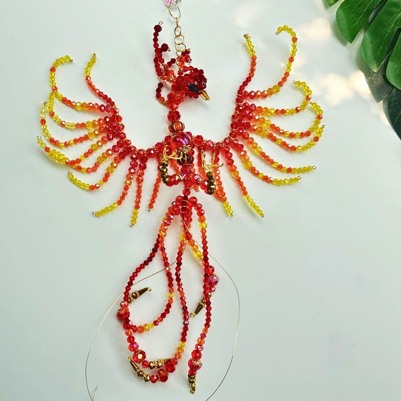 May include: A beaded phoenix sculpture in shades of red, orange, and yellow. The bird has outstretched wings and a long, flowing tail. The sculpture is made of small beads strung together with wire, creating a delicate and detailed design.
