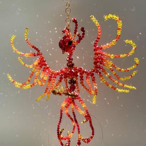 May include: A handcrafted beaded phoenix sculpture in shades of red, orange, and yellow. The bird is suspended by a gold chain, with wings outstretched. The sculpture is made of small, colorful beads, creating a detailed and vibrant design.