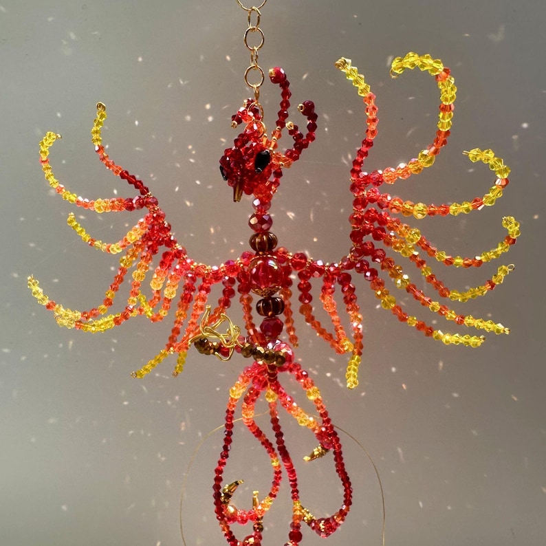 May include: A handcrafted beaded phoenix sculpture in shades of red, orange, and yellow. The bird is suspended by a gold chain, with wings outstretched. The sculpture is made of small, colorful beads, creating a detailed and vibrant design.