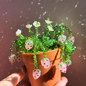 May include: A decorative plant sculpture in a terracotta pot featuring beaded green vines with white flower blossoms and translucent pale pink strawberry charms with red dots. The stems are constructed from faceted green glass beads.
