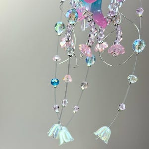 May include: A delicate, handcrafted mobile featuring strands of iridescent beads and small, pink flower-shaped accents. The mobile includes clear, blue, and pink beads, with white, bell-shaped flowers at the bottom. The design is suspended by thin, silver wires.