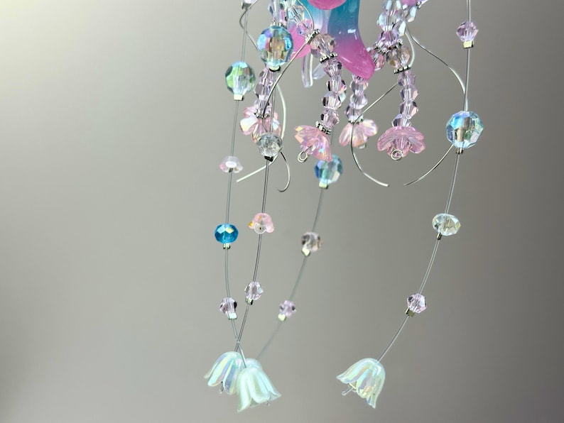 May include: A delicate, handcrafted mobile featuring strands of iridescent beads and small, pink flower-shaped accents. The mobile includes clear, blue, and pink beads, with white, bell-shaped flowers at the bottom. The design is suspended by thin, silver wires.