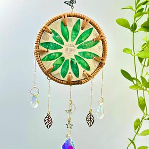 Op de afbeelding: Een decoratieve dromenvanger met een cirkelvormig geweven frame. Het midden heeft een groen bladeren ontwerp. Aan de onderkant hangen kristallen hangers, bladbedels en een maan- en sterbedel. Een vlinderbedel en een kristallen kraal zitten bovenaan.
