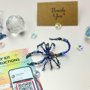 May include: A DIY kit with instructions and a beaded scorpion sculpture. The scorpion is dark blue with gold and iridescent accents. The kit includes a clear crystal ball, a thank you card, and a smartphone with a QR code.