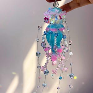 May include: A handcrafted jellyfish ornament with a turquoise body and pink accents. The ornament is adorned with iridescent beads, clear crystals, and delicate wire strands. The top features a light blue and pink design with butterfly accents.