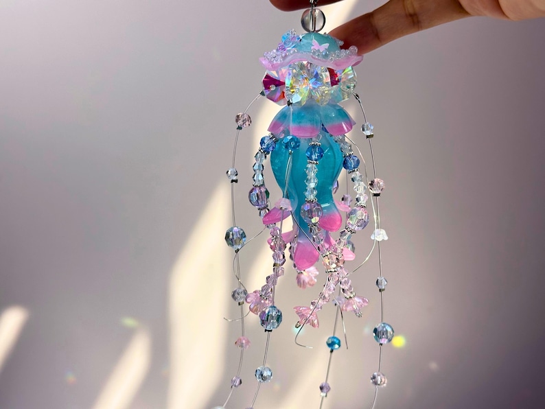 May include: A handcrafted jellyfish ornament with a turquoise body and pink accents. The ornament is adorned with iridescent beads, clear crystals, and delicate wire strands. The top features a light blue and pink design with butterfly accents.