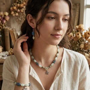 Puede incluir: Un conjunto de joyas con collar, pulsera y pendientes. El collar y la pulsera tienen una mezcla de cuentas verde claro, azules y plateadas, con un colgante de flor plateada. Los pendientes tienen cuentas azules y blancas.