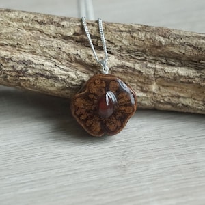 Può includere: Un ciondolo marrone a forma di fiore con una pietra preziosa rossa al centro, appeso a una catena d'argento. Il ciondolo è appoggiato su un pezzo di legno galleggiante.