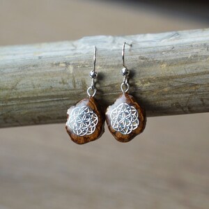 Può includere: Un paio di orecchini d'argento con ciondoli in legno marrone a forma di lacrima con un design geometrico argentato.