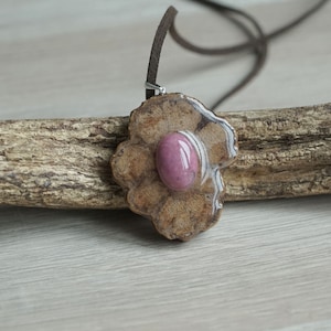Può includere: Un ciondolo in pietra rosa con una montatura in resina marrone, appeso a un cordino in pelle marrone.
