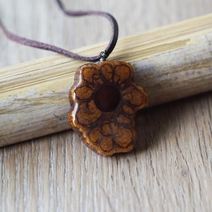 Può includere: Una collana con un ciondolo in ceramica a forma di fiore marrone con un centro marrone scuro e dettagli intricati. La collana è su un cordoncino in pelle marrone.