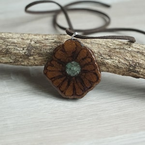 Può includere: Un ciondolo a forma di fiore in ceramica marrone con un centro verde, appeso a un cordino in pelle marrone.