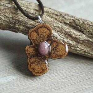 Può includere: Una collana con un ciondolo a forma di fiore in legno con una pietra rosa al centro. Il ciondolo è marrone e presenta un design dettagliato. La collana è realizzata con un cordoncino in camoscio marrone.