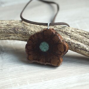 Può includere: Un ciondolo in legno marrone con una pietra verde al centro, appeso a un cordino in pelle marrone.