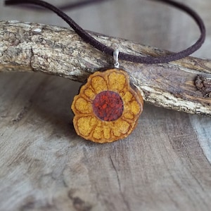 Può includere: Una collana con un ciondolo a forma di fiore in legno con un centro rosso e petali gialli. La collana è realizzata con un cordoncino in camoscio marrone.