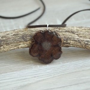 Può includere: Un ciondolo a forma di fiore in ceramica marrone con un centro bianco, appeso a un cordino in pelle marrone.