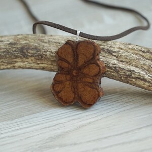 Può includere: Una collana con un ciondolo di legno marrone a forma di fiore, con un cordoncino di cuoio. Il fiore ha un design dettagliato ed è appeso a un pezzo di legno galleggiante.