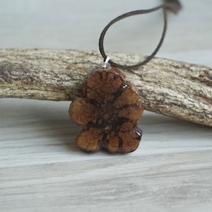 Può includere: Un ciondolo in legno marrone con un motivo unico e intricato appeso a un cordoncino in pelle marrone. Il ciondolo ha la forma di un fiore a cinque petali.