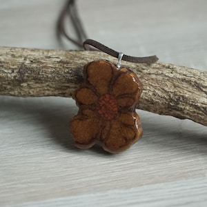 Può includere: Un ciondolo in ceramica marrone a forma di fiore con un centro rosso, appeso a un cordino in pelle marrone.