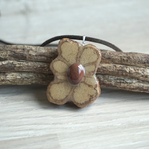 Può includere: Una collana con un ciondolo a forma di fiore in ceramica con una pietra marrone al centro. Il ciondolo è beige con linee marroni ed è appeso a un cordoncino di cuoio marrone.