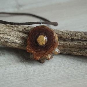 May include: A brown wooden pendant with a circular design and a light brown center. The pendant is hanging from a brown leather cord.