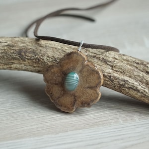 Può includere: Un ciondolo a forma di fiore in ceramica marrone con una pietra di agata verde e bianca al centro, appeso a un cordino di cuoio marrone.