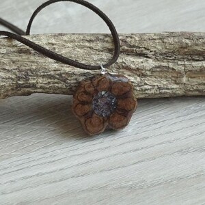 Può includere: Un ciondolo a forma di fiore in legno marrone con un centro viola e bianco, appeso a un cordino in pelle marrone.