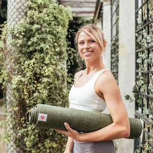 May include: A woman wearing a white sports bra holds a rolled up green yoga mat. She is smiling and looking at the camera.