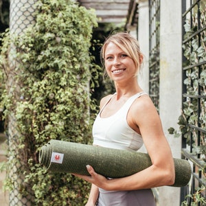 May include: A woman in a white sports bra holds a rolled up green yoga mat. She is smiling and looking at the camera.