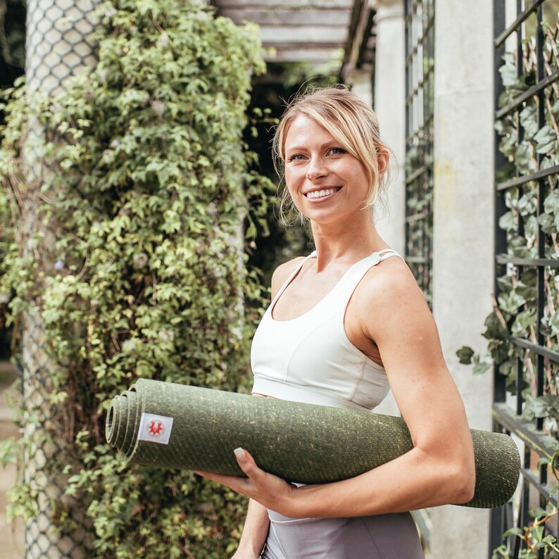 May include: A woman in a white sports bra holds a rolled up green yoga mat. She is smiling and looking at the camera.