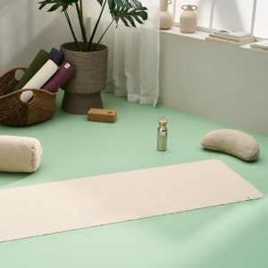 May include: A light beige yoga mat, a brown yoga block, a beige yoga bolster, a silver water bottle, and a beige crescent-shaped yoga cushion on a green surface.