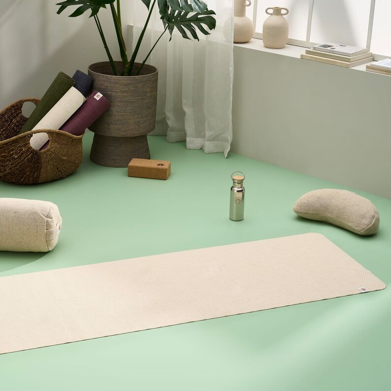 May include: A light beige yoga mat, a brown yoga block, a beige yoga bolster, a silver water bottle, and a beige crescent-shaped yoga cushion on a green surface.