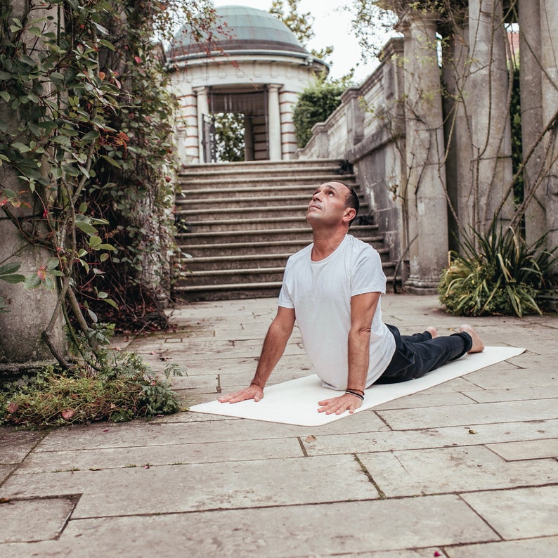 May include: A man wearing a white t-shirt and black trousers is performing a yoga pose on a white mat in a garden setting. He is looking upwards with his arms stretched out in front of him.