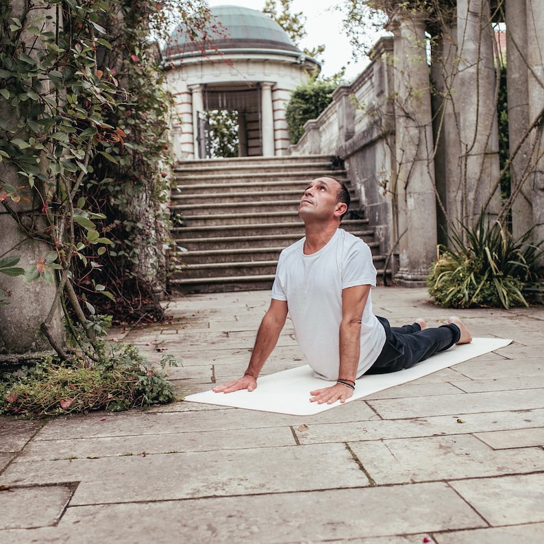 May include: A man in a white t-shirt and black pants is doing a yoga pose on a white mat in a garden setting. He is looking up at the sky with his arms extended forward.