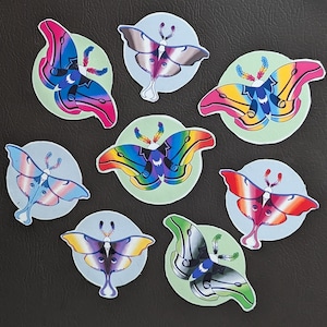May include: A collection of eight moth stickers with various colorful wing designs. Each moth is set on a light green circle, with a light blue background. The moth wings feature shades of pink, blue, yellow, and rainbow patterns.