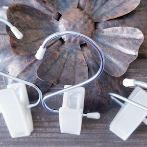 May include: Three white plastic clips with silver metal hooks. The clips are attached to a silver metal wire that forms a loop. The clips are on a wooden surface.