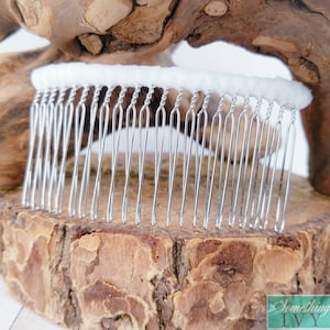 May include: A silver hair comb with a white fabric base. The comb has a row of small, closely spaced teeth.