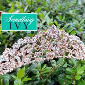 Puede incluir: Una tiara de oro rosa con detalles de cristal transparente, sobre un lecho de hojas verdes. La tiara presenta un dise&ntilde;o ornamentado con piedras brillantes. La imagen incluye un rect&aacute;ngulo turquesa con el texto "Something IVY".