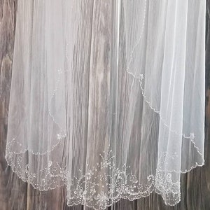 May include: A white bridal veil with a beaded edge and floral pattern. The veil is displayed on a white mannequin against a wood background.