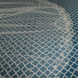 May include: A white bridal veil with a beaded edge. The veil is laid out on a blue and white patterned surface.