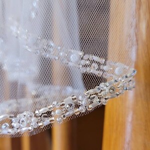 May include: Close-up of a white tulle veil with a beaded trim. The trim features silver beads, clear crystals, and white pearls.