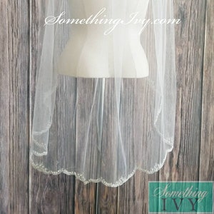May include: A white bridal veil with a scalloped edge and a beaded trim. The veil is displayed on a white mannequin against a wood background.